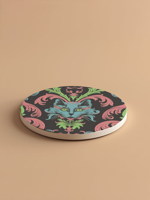 Circular ceramic coaster featuring a turquoise cat face with green eyes on black background, surrounded by pink scrollwork and green leaves in Victorian damask style.